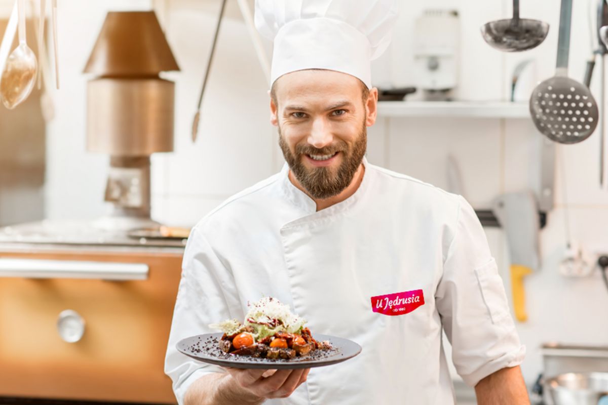 Kucharz prezentujący gotowe danie w restauracyjnej kuchni Kucharz w białym stroju prezentuje talerz z daniem w profesjonalnej kuchni.