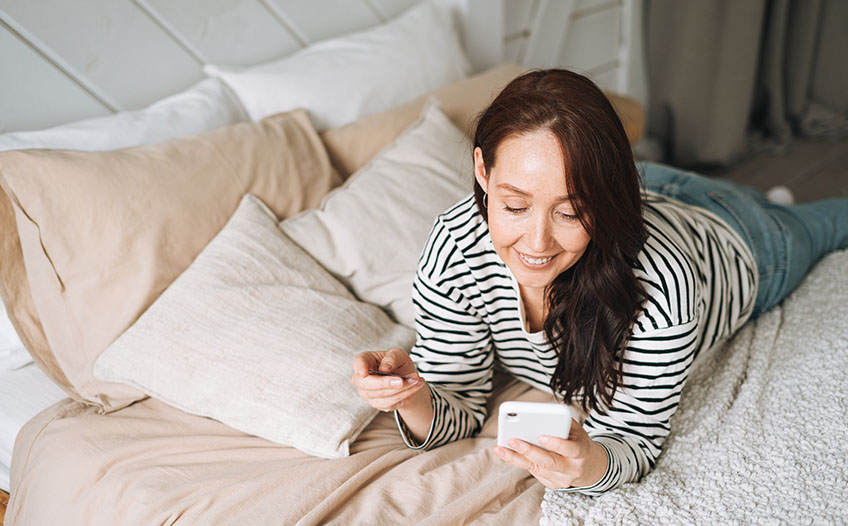 Kobieta leżąca na łóżku buszuje w internecie w smartfonie